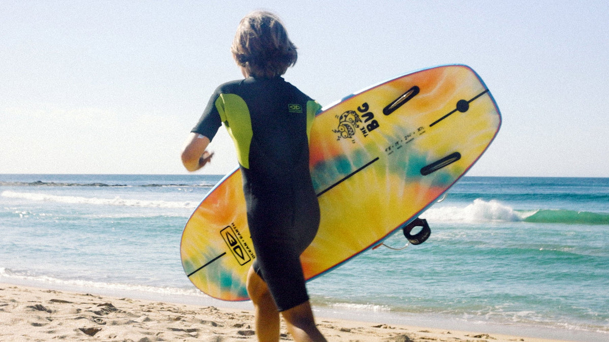 A person on a sandy beach, seen from behind, carrying a brightly colored surfboard under their arm while wearing a black and green wetsuit. The ocean and blue sky are in the background.